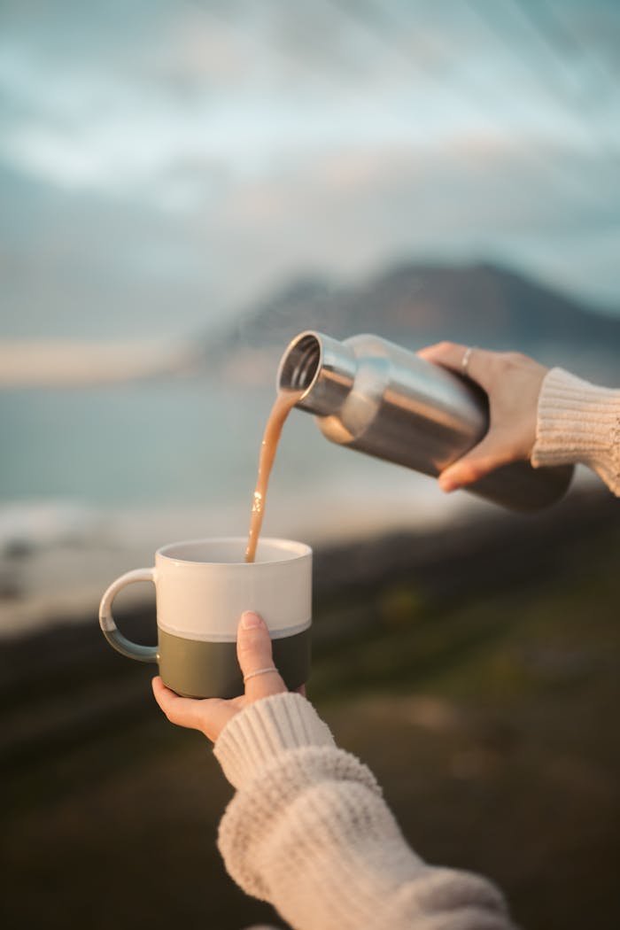 Hands pouring hot beverage from thermos into a cup with mountains in the background.